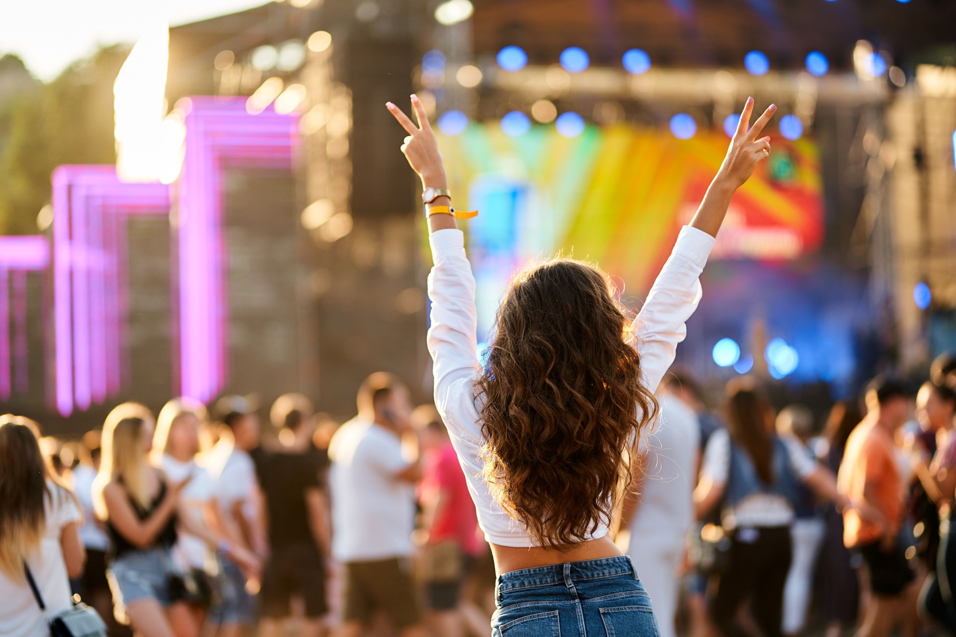 Back view of young woman at summer beach music festival. Raised hands with peace sign, enjoying live concert. Crowd, stage lights, sunset party vibes. Casual fashion, outdoor event, fun atmosphere.