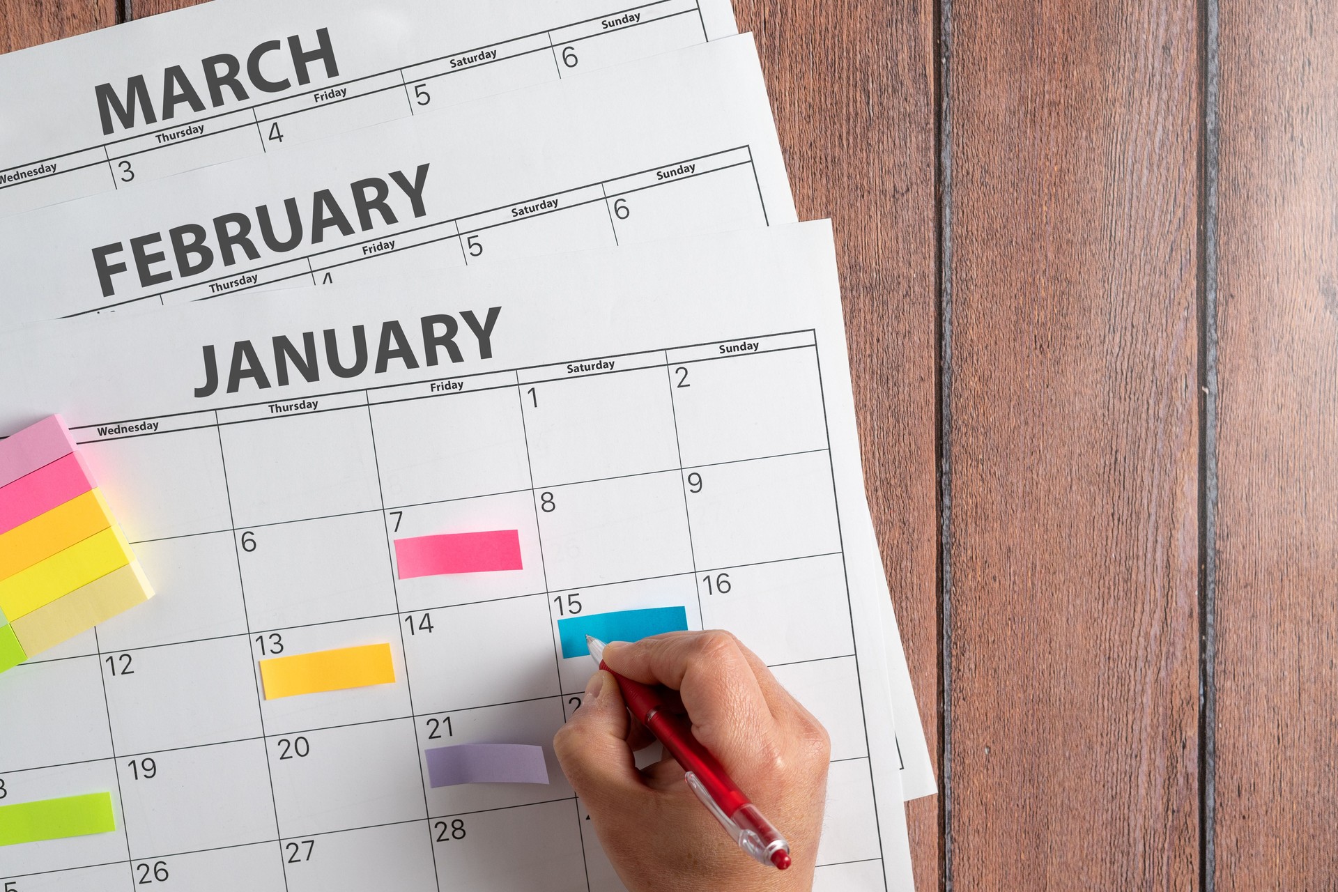 View from above of a person's hand making notes on a calendar with the first months of the year using colored post it notes to highlight the activities. Concept plans and objectives, fulfillment of goals